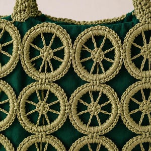 May include: Close-up of a green tote bag with a repeating pattern of crocheted wagon wheel designs in a lighter green. The bag has a dark green fabric base and matching crocheted handles.
