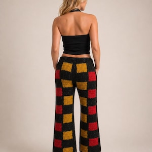 Op de afbeelding: Een zwarte haltertop gecombineerd met een broek met hoge taille en wijde pijpen. De broek heeft een dambordpatroon in zwart, rood en goud. De outfit wordt gecompleteerd met zwarte sandalen met hakken. De algehele stijl is retro en bohemien.