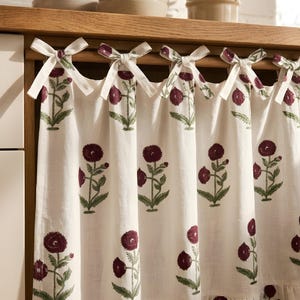 Peut inclure: Un rideau en tissu blanc orné d'un motif floral répétitif composé de fleurs marron foncé avec des tiges et des feuilles vertes. Le rideau est fixé à une tringle en bois par des nœuds en tissu et présente un ourlet à volants en bas.