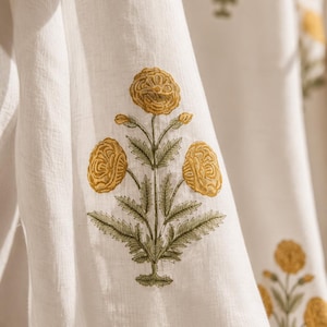 May include: Close-up of white fabric with a repeating floral pattern. The design features yellow flowers with detailed petals and green leaves. The fabric has a soft, textured appearance, suggesting a natural material.