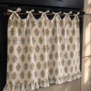 May include: White kitchen curtain with a repeating floral design in yellow and green. The curtain has a frilled edge and is tied to a rod with small bows. The curtain is hanging in front of a black oven.