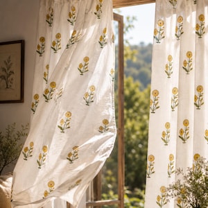 Peut inclure: Rideaux en lin blanc ornés d'un motif floral jaune répété. Les rideaux sont suspendus à une tringle en bois, la lumière du soleil filtrant à travers une fenêtre ouverte. Les rideaux sont attachés en haut avec du tissu blanc.