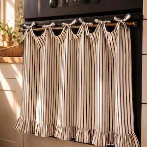 May include: A kitchen curtain featuring a vertical dark brown and cream striped pattern. The fabric hangs from a rod with six tied fabric bows at the top and includes a gathered ruffle trim along the bottom edge, covering the front of a black oven.