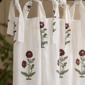 May include: White cotton curtains with a repeating floral pattern in burgundy and green. The curtains are tied to a black rod with matching white fabric ties, creating a cohesive and decorative window treatment.
