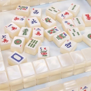 May include: A collection of off-white Mahjong tiles featuring various engraved symbols, characters, and numbers in red, green, and blue ink. The tiles are arranged in a mix of scattered pieces and organized rows, showcasing a smooth, glossy finish.