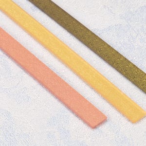 May include: Three long, thin, rectangular strips arranged diagonally. The strips are colored in shades of light coral, pale yellow, and dark bronze. Each strip features a fine, textured surface finish that appears slightly granular.