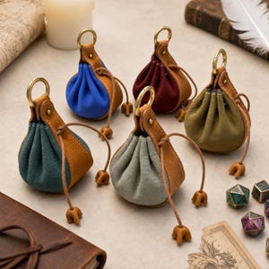 May include: Five small leather drawstring pouches in blue, dark red, olive green, teal, and light grey. Each pouch features a tan leather top section, a gold-toned metal ring at the top, and a matching tan leather drawstring with small decorative ends.
