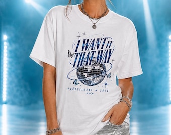 Pode incluir: Uma t-shirt branca de manga curta com uma estampa gráfica azul com o texto I WANT IT THAT WAY, uma bola de discoteca e borboletas. Abaixo do gráfico, o texto diz DÜSSELDORF 2026. A t-shirt é combinada com calções de ganga prateados metalizados.