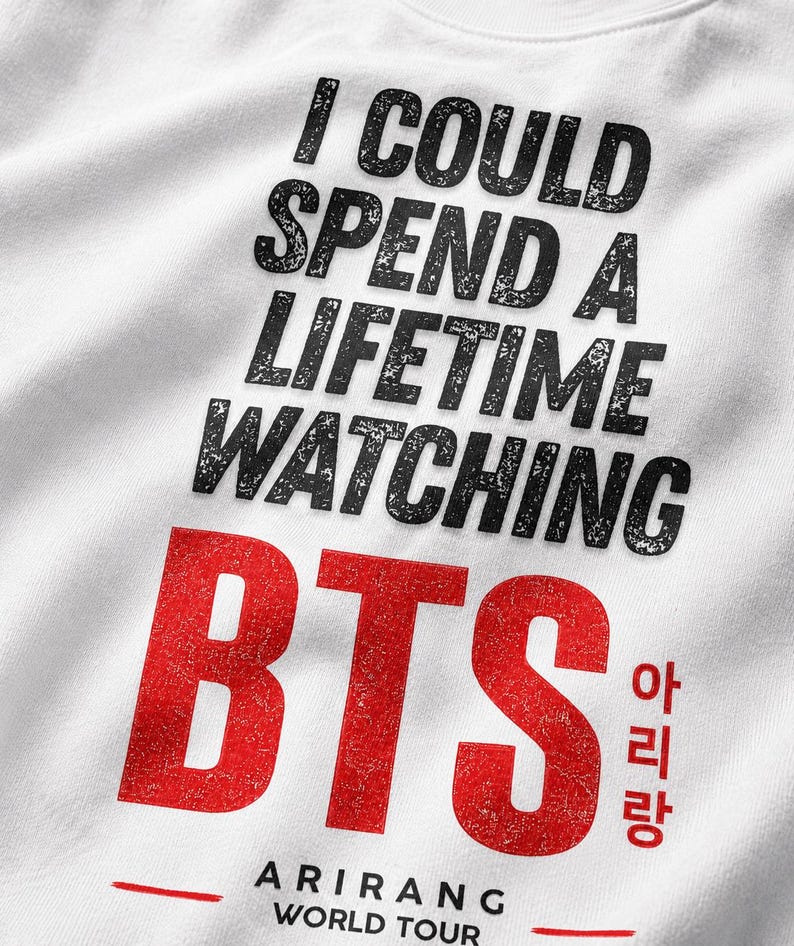 Peut inclure: Sweat-shirt blanc avec l'inscription "I COULD SPEND A LIFETIME WATCHING BTS" en noir, avec un effet vieilli. Le mot "BTS" est en grandes lettres rouges, &eacute;galement avec un effet vieilli. En dessous, le texte "ARIRANG WORLD TOUR" est imprim&eacute; en rouge.