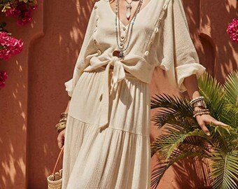 May include: A cream-colored two-piece set consisting of a V-neck blouse with long sleeves, shell accents, and a front tie, paired with a matching tiered maxi skirt. The outfit is accessorized with layered necklaces, hoop earrings, and bracelets.