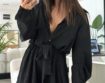 May include: A black long-sleeved mini dress with a textured, crinkled fabric finish. The dress features a collared neckline, a button-down front, and a matching fabric tie belt at the waist. The sleeves are long and loose with gathered cuffs.