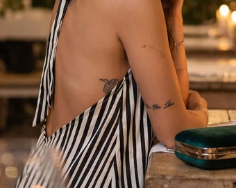 May include: A black and white striped halter top dress with a tie-neck closure and an open back design. The garment features vertical stripes throughout and is paired with a small, rectangular, dark green velvet clutch bag.