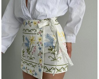 May include: A white long-sleeved button-down shirt paired with a white wrap skirt featuring colorful floral embroidery. The skirt has a tie-waist closure and is decorated with green, yellow, and blue botanical patterns arranged in horizontal bands.
