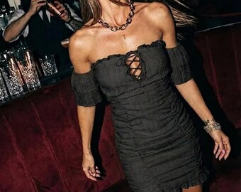 May include: A black off-the-shoulder mini dress featuring a ruched texture, short sleeves, and a lace-up detail at the center of the chest. The garment has a ruffled hemline and a fitted silhouette, crafted from a lightweight fabric.