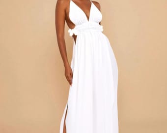 May include: A floor-length white maxi dress featuring a deep V-neckline, thin spaghetti straps, and a cutout waist section with ruffled detailing. The skirt includes a high side slit, and the fabric has a flowing, lightweight appearance.