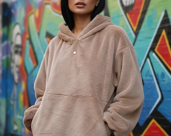 May include: A beige, long-sleeved, plush faux-fur hoodie featuring a large front kangaroo pocket, a hood, and a relaxed fit. The garment is made of a soft, textured material and is worn over white trousers, with a simple gold pendant necklace.