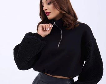 May include: A black cropped sweatshirt featuring a high collar with a silver-toned zipper closure. The garment has long, voluminous sleeves made of a textured, fuzzy fleece material, while the bodice is constructed from a smooth fabric.