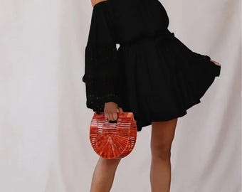 May include: A black off-the-shoulder dress with long sleeves featuring eyelet lace detailing, paired with a semi-circular orange wooden handbag and black wedge sandals with ankle ties and woven jute platforms.