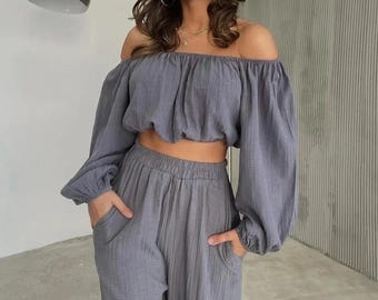 May include: A two-piece set in a muted gray color featuring a cropped off-the-shoulder top with long, billowy sleeves and matching high-waisted trousers with side pockets. The fabric has a visible crinkled texture throughout the garment.