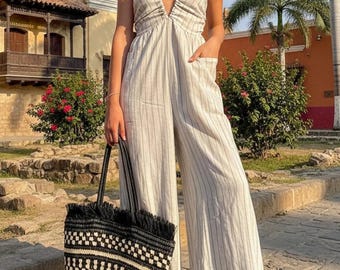 May include: A beige jumpsuit with thin vertical dark stripes, a halter neckline, and side pockets, paired with a large black and white woven tote bag featuring a fringed top edge and a geometric pattern, worn with simple flat sandals.