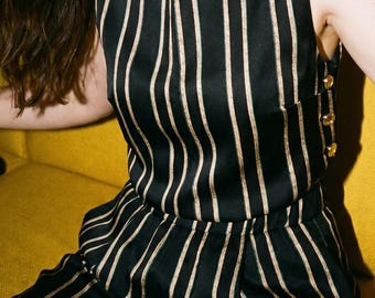 May include: A sleeveless black top and matching trousers set featuring vertical gold stripes. The top has a high neckline with gold buttons along the shoulder and side seam. The trousers have a high-waisted fit with a pleated front design.