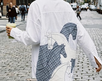 May include: A white long-sleeved button-down shirt featuring a large back panel with embroidered horse silhouettes. The horse designs are filled with a dark blue and white textured pattern, contrasting against the plain white fabric of the shirt.