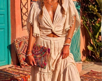 May include: A beige two-piece set consisting of a short-sleeved top with a front twist detail and a long, tiered skirt. The top and skirt feature small white shell trim along the edges. A colorful, patterned clutch with fringe is held in one hand.