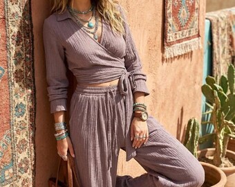 May include: A mauve two-piece set consisting of a long-sleeved wrap top with a side tie and matching wide-leg trousers with an elastic drawstring waist. The outfit is made of a textured fabric and is paired with a straw hat and a brown leather tote bag.