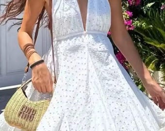 May include: A white halter-neck mini dress featuring an all-over eyelet embroidery pattern with a deep V-neckline and a flared skirt, paired with a small, tan woven straw handbag with a prominent gold-toned logo on the front.