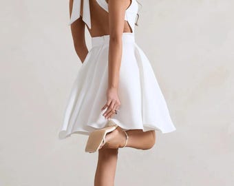 May include: A white mini dress featuring a sleeveless bodice, a high neckline, and a large bow detail at the back of the neck. The dress has an open back design, a fitted waistline, and a flared, A-line skirt that falls above the knees.