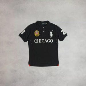 Puede incluir: Polo negro con texto "CHICAGO" en blanco y un logotipo de jugador de polo. Presenta el número "3" en la manga y un escudo. Acentos rojos en el dobladillo. Un diseño clásico y deportivo.