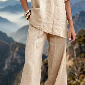 May include: A beige linen two-piece outfit, featuring a V-neck top with short sleeves and wide-leg pants. The outfit is made of a lightweight fabric, perfect for warm weather. The model is wearing gold bracelets and burgundy heels.