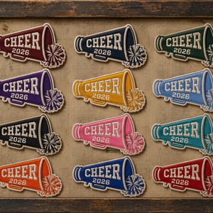 May include: A collection of twelve megaphone-shaped patches arranged in a grid. Each patch features the word CHEER and the year 2026 in white lettering, accompanied by a matching pom-pom graphic. The patches come in various colors including red, navy, green, purple, yellow, light blue, black, pink, teal, orange, and royal blue.