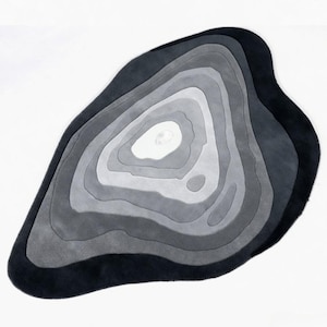Könnte beinhalten: Abstrakter Teppich in Grautönen, der an einen geologischen Querschnitt erinnert. Die konzentrischen Schichten wechseln von einem dunklen Kohlefaserrand zu einem hellen weißen Zentrum und bilden eine organische Form.