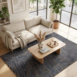 Könnte beinhalten: Ein rechteckiger Teppich mit einem geometrischen Linienmuster in Dunkelblau und Beige. Der Teppich liegt auf einem hellen Holzboden unter einem beigen Sofa, einem Couchtisch aus Holz, einer Keramikvase mit Trockengras und einem Holztablett.