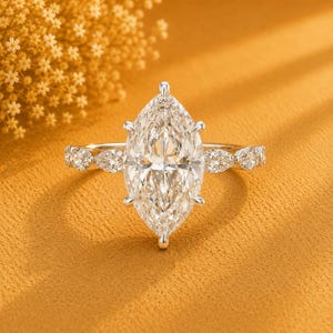 Op de afbeelding: Een zilverkleurige ring met een grote marquise-geslepen centrale steen die door zes pootjes wordt vastgehouden. De band is versierd met een reeks kleinere, marquise- en rondgeslepen stenen die symmetrisch aan beide zijden van de zetting zitten.