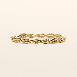 Puede incluir: Un anillo de oro con un diseño trenzado que combina una banda lisa y pulida con un hilo más fino de textura similar a una cuerda. Ambos elementos están entrelazados para formar un patrón circular continuo en oro amarillo brillante.