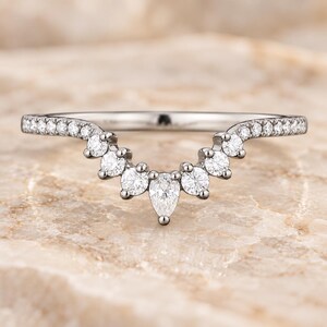 May include: A silver-toned curved ring featuring a central pear-cut stone flanked by round-cut stones, with additional small round stones set along the band. The metal band follows a V-shaped contour, creating a delicate and symmetrical design.