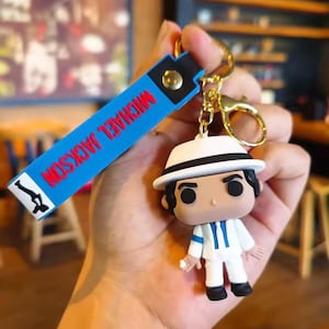 May include: A Michael Jackson keychain featuring a Funko Pop figure in a white suit and fedora. The blue strap reads "MICHAEL JACKSON" in red letters. The keychain has a gold-colored clasp and a small, white object in the figure's hand.