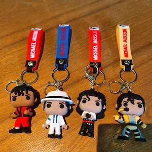 May include: Four keychains featuring Michael Jackson figures in various outfits. Each keychain has a strap with "MICHAEL JACKSON" printed on it. The straps are red, blue, red, and yellow. The figures are in red, white, black, and yellow outfits.