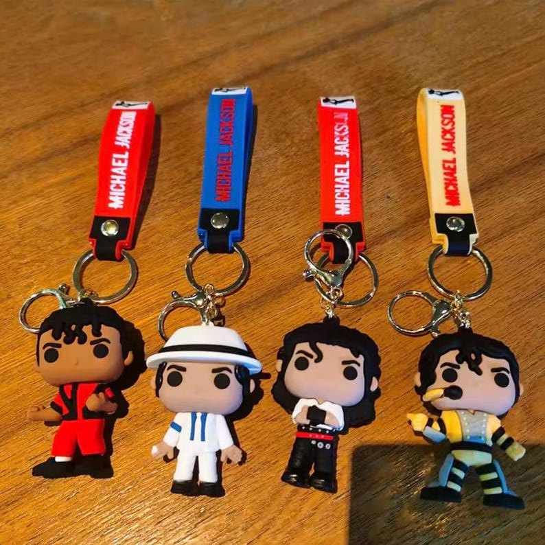 May include: Four keychains featuring Michael Jackson figures in various outfits. Each keychain has a strap with "MICHAEL JACKSON" printed on it. The straps are red, blue, red, and yellow. The figures are in red, white, black, and yellow outfits.