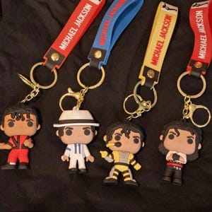 May include: Four keychains featuring Michael Jackson figures in various outfits. Each keychain has a strap with "MICHAEL JACKSON" printed on it. The straps are red, blue, and yellow. The figures are in red, white, yellow, and black outfits.