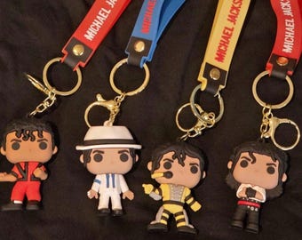 May include: Four keychains featuring Michael Jackson figures in various outfits. Each keychain has a strap with "MICHAEL JACKSON" printed on it. The straps are red, blue, and yellow. The figures are in red, white, yellow, and black outfits.