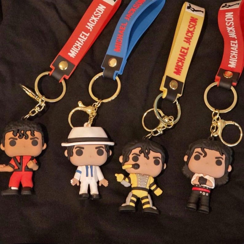 May include: Four keychains featuring Michael Jackson figures in various outfits. Each keychain has a strap with "MICHAEL JACKSON" printed on it. The straps are red, blue, and yellow. The figures are in red, white, yellow, and black outfits.
