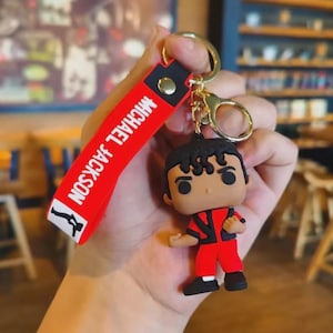 May include: A red and black keychain featuring a Michael Jackson figure. The figure is wearing a red outfit with black accents. The keychain strap is red with "MICHAEL JACKSON" printed in white.