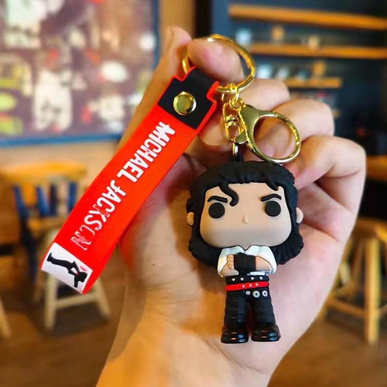 May include: A Michael Jackson keychain featuring a stylized figure with black hair, a white shirt, and black pants. The red strap reads "MICHAEL JACKSON" in white letters. The keychain has a gold-colored clasp.