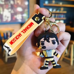 May include: A keychain featuring a Michael Jackson figurine in a yellow and black outfit, with a microphone. The keychain strap is yellow with the text "MICHAEL JACKSON" in red. The keychain has a gold-colored clasp.