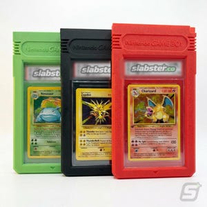 May include: Three graded Pokémon cards in protective cases. The cases are green, black, and red, resembling Game Boy cartridges. Each case has the text "Nintendo GAME BOY" and "slabster.co". The cards feature characters like Venusaur, Zapdos, and Charizard.