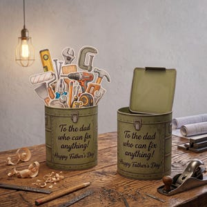 Puede incluir: Dos cajas de herramientas de metal verde oliva con el texto "To the dad who can fix anything! Happy Father's Day!". Una caja de herramientas está abierta y la otra contiene recortes de herramientas. La escena está ambientada sobre una superficie de madera.