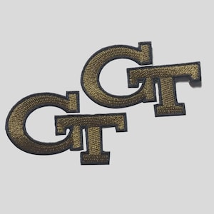 May include: Two embroidered patches featuring the letters GT in a stylized font. The letters are gold with a dark navy blue border. The embroidery has a textured, raised appearance with visible stitching throughout the design of the letters.